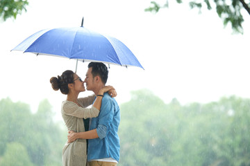 Side view portrait of affectionate Asian couple embracing under umbrella in rain, copy space