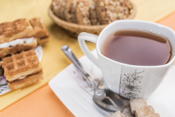 White mug with tea, a spoon and lump brown sugar lie on a white square saucer, next to the defocus of Viennese waffles and cookies on a warm pear-yellow and light orange background.