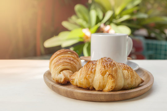 Fresh croissant and hot coffee on round wooden plate with morning warm light over tropical garden background