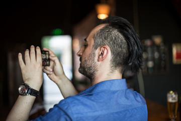 Fototapeta premium Young bearded man taking photos with his mobile phone in the bar