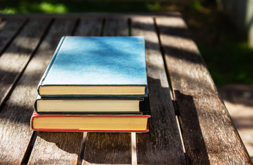 Three books on top of a wooden table, outside. This image can be used represent reading or education.