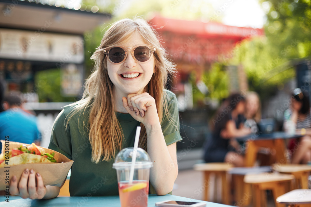 Wall mural Portrait of a blonde freckled girl cheerfully smiling holding a taco sitting at a table on a food cort in park during lunch on a summer day.