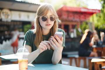 Portrait of young blonde woman writing a blog post on her pink cell phone or chatting with friends in social media wearing glasses on a sunny day in park.