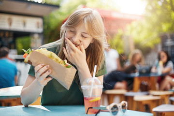 Girl eating taco smiling. Hungry freckled blonde woman eating junk food on a food court drinking lemonade on a sunny summer day in park.