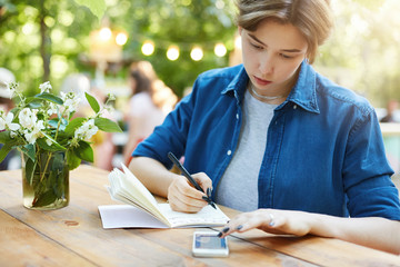 Girl taking notes using smartphone. Outdoor portrait of a young woman writing in her notebook preparing for exam or planning winter holidays.