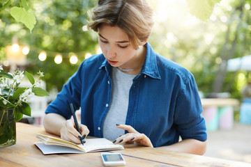 Woman taking notes in notepad. Outdoor portrait of a young girl planning her vacation or a new startup business using a pen and a smartphone in park in summer wearing blue shirt.