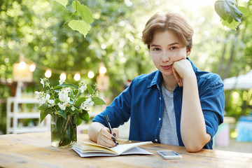 Girl preparing for exam. Outdoor portrait of a confident young woman writing in notepad and looking at camera preparing for her study or work. Soon to become a scriptwriter.