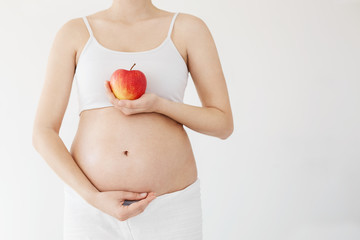 Front portrait of healthy pregnant woman holding her belly and red apple full of vitamins. Pregnancy healthy diet concept over white. High key.