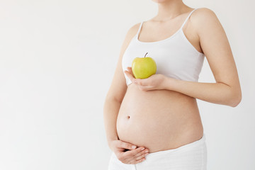 Pregnant woman holding her tummy and a healthy green apple full of vitamins waiting for her baby. Prenatal diet concept. High key.