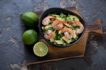 Salad with avocado, cucumbers and tiger shrimps served in a bowl, studio shot on a brown stone background