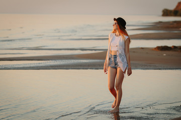 Young, beautiful girl walking at the beach at sunset. Stylish woman with long hair standing in blouse and jeans shorts