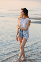 Young, beautiful girl walking at the beach at sunset. Stylish woman with long hair standing in blouse and jeans shorts