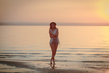 Young, beautiful girl walking at the beach at sunset. Stylish woman with long hair standing in blouse and jeans shorts