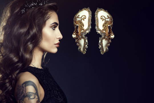 Close up portrait of charming lady with dark wavy silky hair and perfect make-up wearing jewel crown and looking in the face of Venetian mask hanging in the air. Backlight. Copy-space. Studio shot