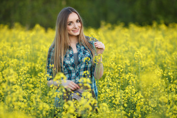 Naklejka na ściany i meble Attractive young blonde woman in blue plaid shirt straw hat enjoying her time on bright colorful blossoming yellow green meadow hard noon sun.