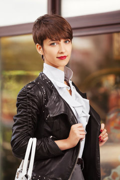 Happy young fashion woman in leather jacket with handbag next to mall window