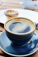 Black coffee with a Portuguese tart in the background 