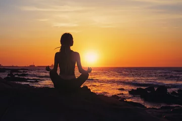 Poster Im Rahmen Silhouette young woman sitting in lotus pose, practicing yoga on ocean beach at sunset © Annatamila