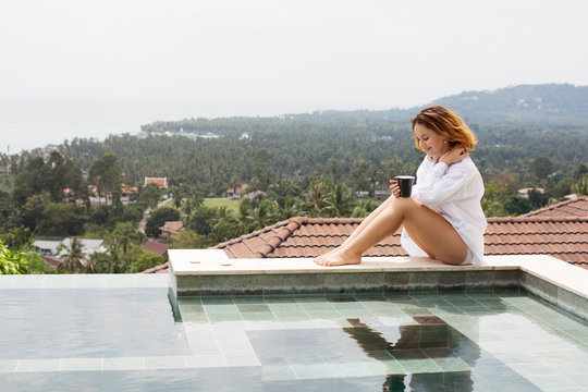 Girl in white shirt with cup of morning coffee sitting near swimming pool of  luxury villa with tropical sea and mountains background