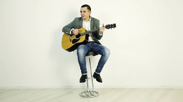 A young guy in a jacket sits on a bar stool and plays an acoustic guitar.