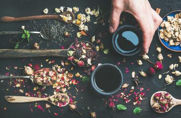 Chinese black tea in black stoneware cups, man's hand holding one cup and wooden spoons with herbs, flower buds and leaves over black wooden background, top view