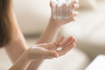 Close up photo of one round white pill in young female hand. Woman takes medicines with glass of...