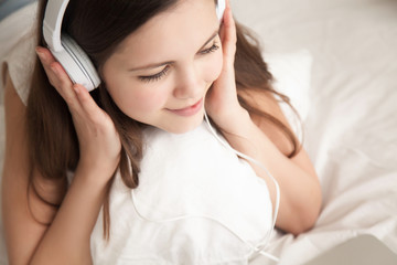 Close up portrait of charming young woman in headphones listening pleasant music while lying in bed with laptop.  Attractive teen girl enjoys internet video, watching online movie, studying distantly