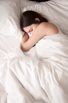 Tired young woman sleeping well on the side in bed with white sheets, resting after sleepless night. Female student lying in bed until late morning. Getting enough sleep concept. View from above
