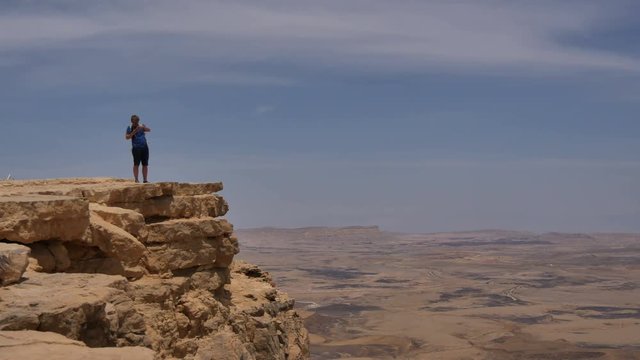 Young man standing on cliff edge and taking pictures of the desert on his phone