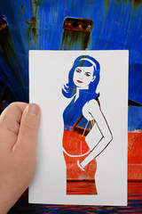 A hand holds a paper stencil of young long-haired girl against background ofblue red boat