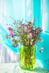 A bouquet of field dry flowers in a vase against a background of gentle air curtains. Natural soft sunlight. Selective soft focus.
