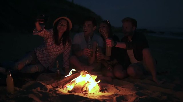 Picnic of young people with bonfire on the beach in the evening. Cheerful friends taking pictures on the phone. Slowmotion shot