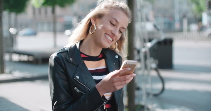 Beautiful young woman wearing leather jacket typing on phone during sunny day.