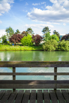 A wooden pontoon with wooden handrail in front of a river with trees and vegetation under a blue sky with white clouds.