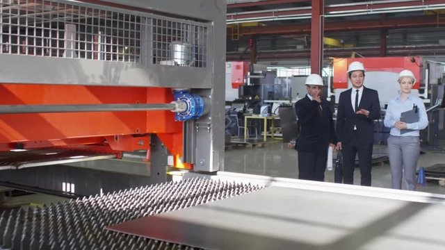 Male and female engineers standing in formal suits and hardhats and listening to their colleague explaining something about machine on factory