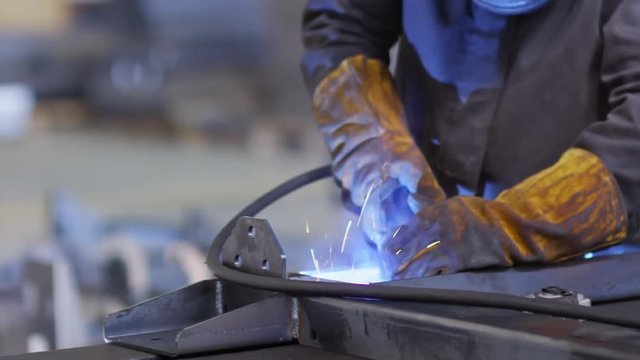 Tracking shot of hands of factory worker in protective gloves welding metal with torch