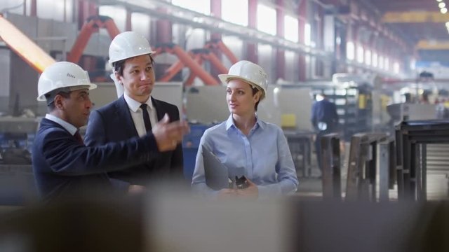 Asian and Latin-American engineers standing with female colleague in factory workshop and discussing something. Woman holding documents and portable radio set