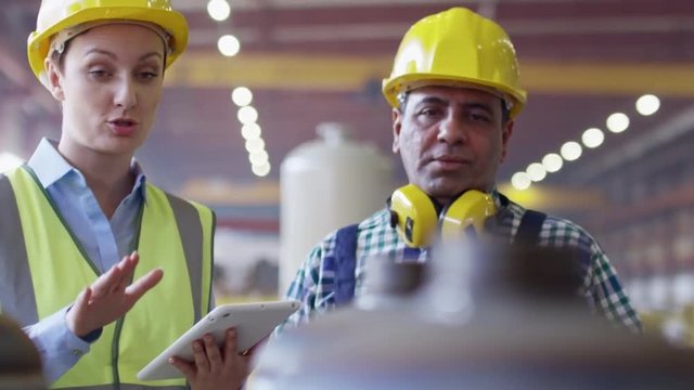 Female factory inspector in uniform and hardhats using digital tablet and talking to male colleague 