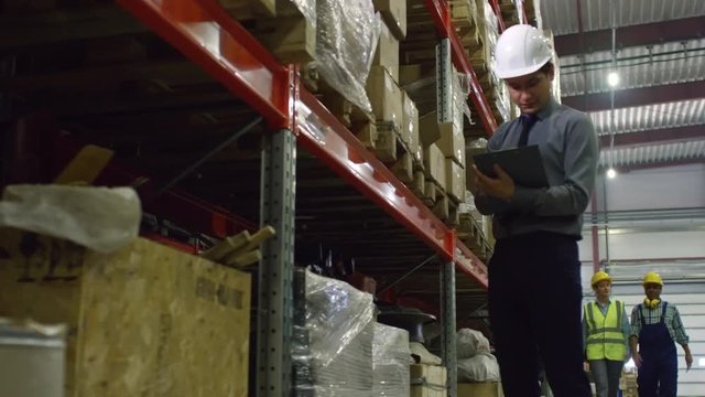 Asian supervisor standing in hardhat in factory warehouse and checking boxes on shelves. He making notes while female technician walking with male colleague behind him