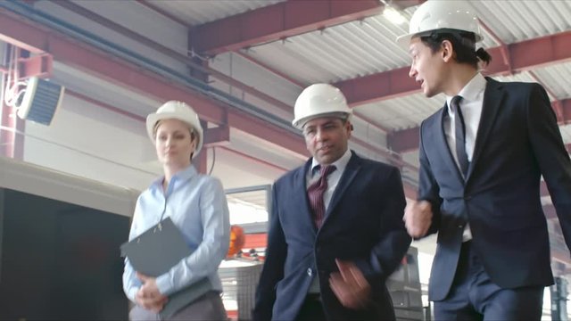 Low angle of two male engineers and woman in hardhat walking in factory and talking about something