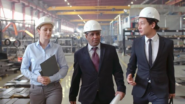 Slow motion shot of Latin American executive engineer in hardhat walking in factory towards camera and talking with male and female colleagues