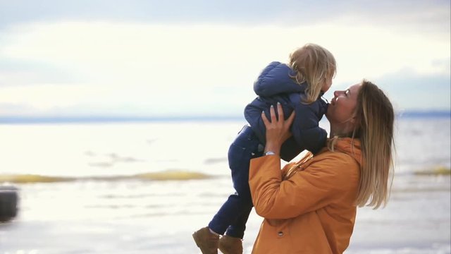 A young mother in a winter jacket plays with her little child on the beach, she throws her son up near the sea, the family enjoys the time together