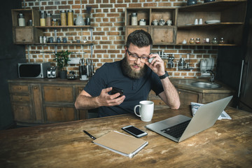 bearded man in eyeglasses sitting at wooden table with laptop, notepad and smartphone in kitchen