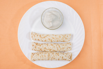 Three pancakes roll with stuffing and sour cream in a bowl in a large white plate on a delicate...