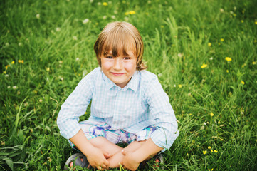 Fototapeta premium Outdoor portrait of adorable 6 year old boy wearing blue shirt, sitting on the grass, top view