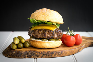 large thick grilled beef minced burger patty with toasted bun melted cheddar cheese green fresh salad cherry tomato and pickle cucumber on rustic wooden board and white table