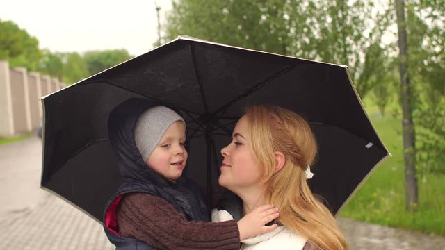 Happy mother holding her little son under an umbrella in a rainy day. The son hugs and kisses mom under an umbrella in the rain and showing thumbs up.