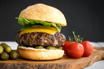 large thick grilled beef minced burger patty with toasted bun melted cheddar cheese green fresh salad cherry tomato and pickle cucumber on rustic wooden board and white table