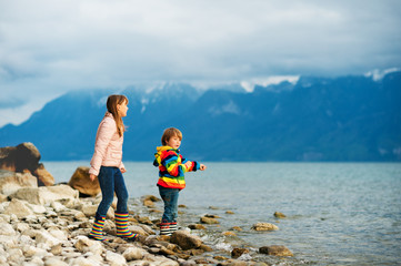 Two happy kids, little brother and big sister, playing together by lake Geneva on a very cloudy day with swiss mountains Alps on background. Image taken in Lausanne, Switzerland