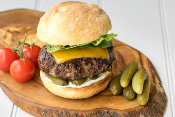large thick grilled beef minced burger patty with toasted bun melted cheddar cheese green fresh salad cherry tomato and pickle cucumber on rustic wooden board and white table